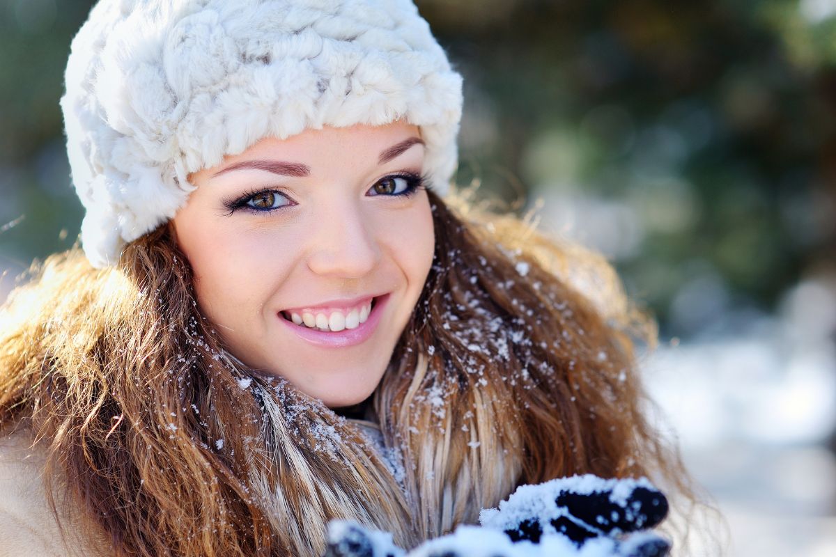 Cold Temperatures and Your Teeth Young woman wearing white knit hat smiling outdoors in snowy winter setting.