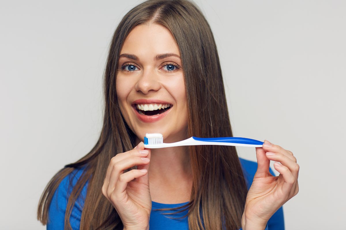 Does My Toothbrush Have Germs Woman in blue shirt holding toothbrush, smiling against neutral background.